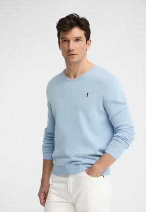 Mann trägt hellblauen V-Ausschnitt-Pullover mit kleinem Golfer-Logo, kombiniert mit weißer Hose, posiert mit einer Hand in der Tasche vor weißem Hintergrund.