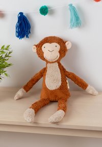 Brown plush monkey toy with a cream belly and hands, soft texture, long limbs, and a smiling face. Sits against a light-colored wall.