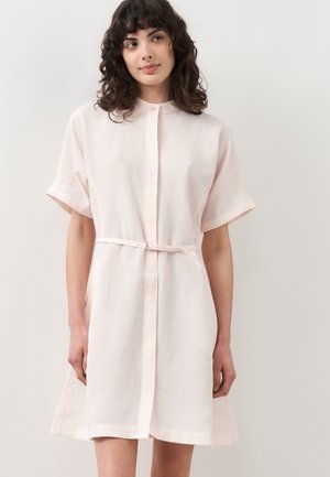 Νεαρή γυναίκα με σγουρά σκούρα μαλλιά που φοράει ένα ανοιχτορόζ φόρεμα με κοντά μανίκια και ζώνη, στέκεται μπροστά σε ένα απλό λευκό φόντο.