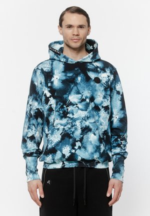 Mężczyzna ubrany w niebiesko-czarną bluzę z abstrakcyjnym wzorem, z założonym kapturem oraz czarne spodnie, stojący na jednolitym białym tle.