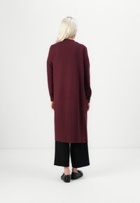 Lange bordeauxrode cardigan met lange mouwen, rechte snit, zijzakken, zachte textuur, gecombineerd met zwarte wijde broek en eenvoudige zwarte schoenen.