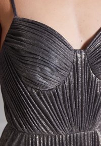 Metaliczna szara sukienka z pionowymi fałdami, szerokim dekoltem i cienkimi ramiączkami. Teksturowany materiał migocze błyszczącym wykończeniem.