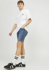 Camiseta de algodón blanca con logo, pantalones cortos de mezclilla con puños doblados, zapatillas negras con rayas blancas y calcetines a rayas en blanco y negro.