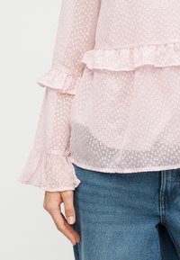 Blouse rose clair texturée avec poignets et détail de taille à volants, portée avec un jean denim bleu, montrant le bas du torse et la main gauche détendue.