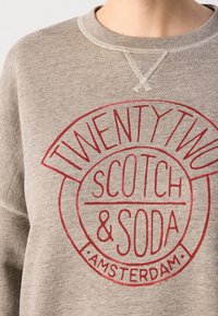 Sudadera gris de algodón con un logo circular rojo que lleva impresa la frase "TWENTY TWO SCOTCH & SODA AMSTERDAM" en una tipografía divertida.