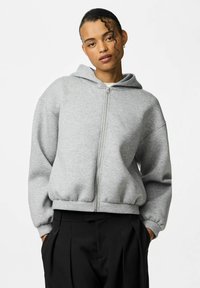 Felpa grigia con zip realizzata in tessuto morbido, dal fit rilassato, con cappuccio ampio e polsini a costine, abbinata a pantaloni neri a vita alta.