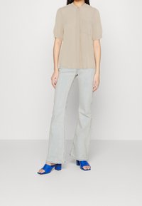 Blusa beige a maniche corte con colletto abbottonato e taschino, abbinata a jeans svasati di un leggero blu e sandali blu con tacco a blocco.