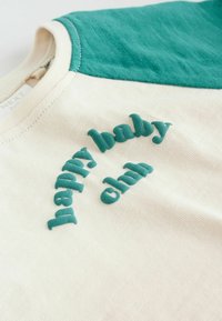 Babyshirt in Creme und Blaugrün mit strukturierten Buchstaben, die nahe dem Kragen vorne "happy baby ohh" lesen.
