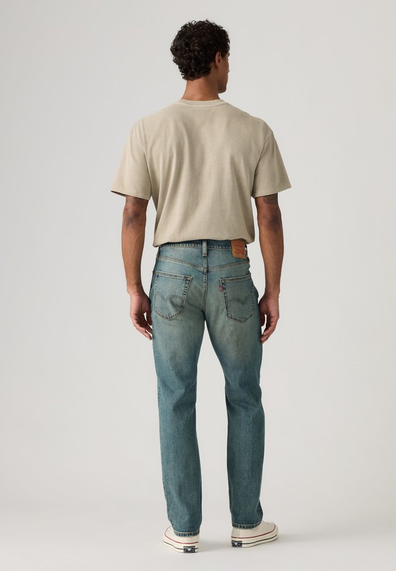 Mand iført lyse beige t-shirt, falmede blå jeans og hvide sneakers, stående med ryggen mod en ensfarvet baggrund.