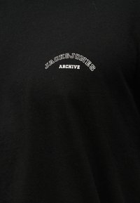 Μαύρο ύφασμα με λευκά γράμματα "JACK & JONES" σε καμπύλη επάνω από τη λέξη "ARCHIVE" στο κέντρο της θωρακικής περιοχής.