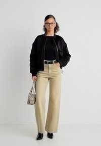 Γυναίκα με μαύρο μπουφάν bomber, μαύρο τοπ, μπεζ παντελόνι ψηλόμεσο με φαρδιά μπατζάκια, μαύρες μπότες, γυαλιά και κρατά τσάντα με σχέδιο φιδιού.