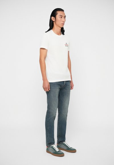 Valkoinen puuvillainen t-paita lyhyillä hihoilla, jossa on pieni punainen kolmion logo rinnassa. Yhdistetty sinisiin farkkuihin ja vihreisiin tennareihin.