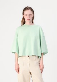 Majica kratkih rukava od pamučne tkanine u mint zelenoj boji, s labavim skraćenim krojem i okruglim izrezom, u kombinaciji s svijetlim bež hlačama. Glatka tekstura, bez uzoraka.