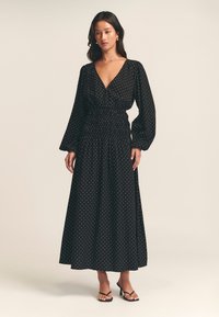 Black maxi dress with white polka dots, deep V-neck, long puff sleeves, smocked waist, and flowing skirt. Worn with black strappy sandals.