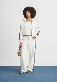 Λευκό κοντό φούτερ με μαύρες ρίγες και λογότυπο, ταιριαστό λευκό παντελόνι φαρδιάς γραμμής με πλαϊνές ρίγες και μικρή τσάντα. Γκρι παπούτσια.