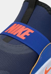 Blå atletisk sko med en teksturert overdel, elastisk stropp med "NIKE" i klar oransje, og kontrasterende oransje detaljer på sålen.