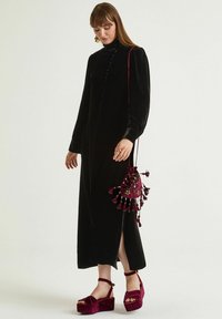 Abito lungo in velluto nero con collo alto e dettagli con bottoni. Abbinato a una borsa con nappine color bordeaux e sandali con zeppa coordinati.