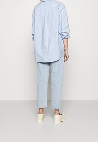 Jasnoniebieska oversize koszula z długimi rękawami, zapięciem na guziki i zaokrąglonym dołem, zestawiona z jasnymi dżinsami i kremowymi trampkami.