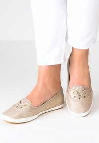 Des chaussures slip-on en toile beige clair présentent des détails en dentelle avec deux œillets dorés, une semelle en caoutchouc blanc et une finition lisse et texturée.