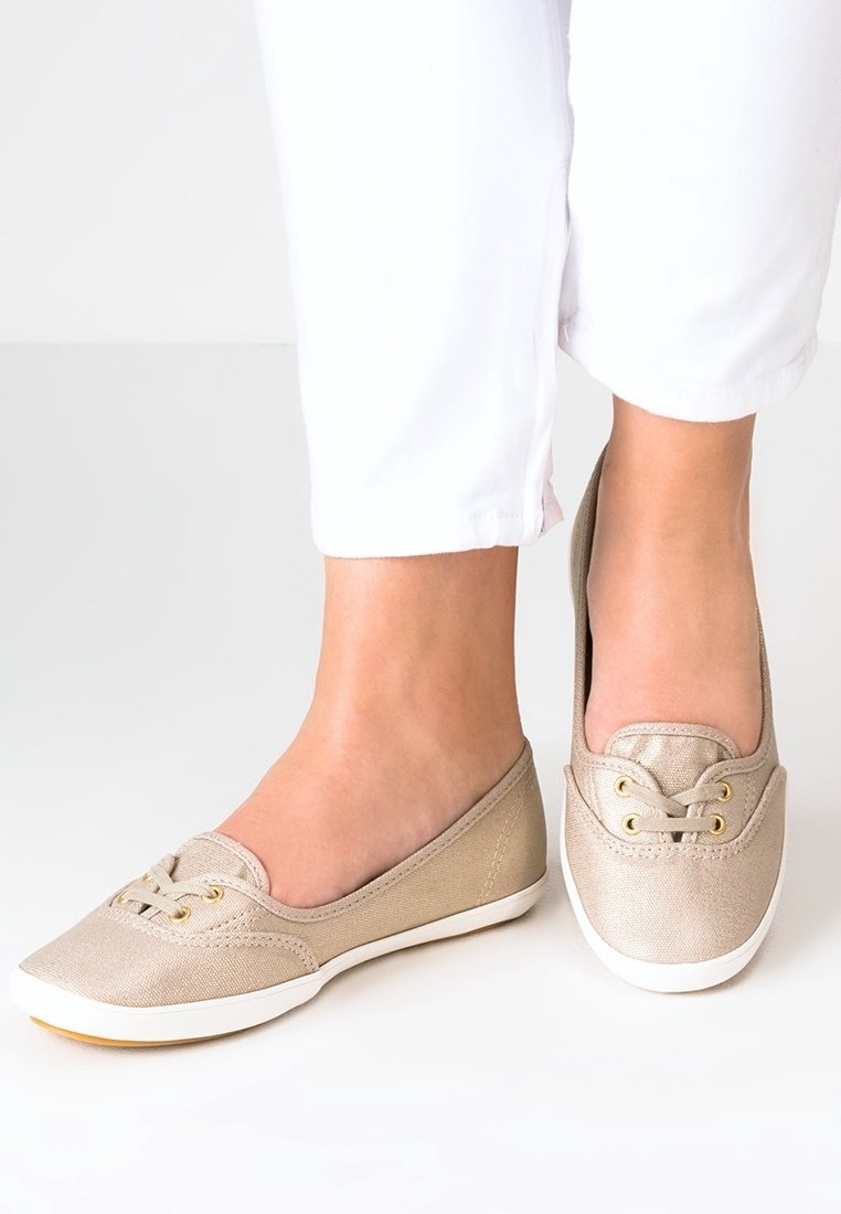 Des chaussures slip-on en toile beige clair présentent des détails en dentelle avec deux œillets dorés, une semelle en caoutchouc blanc et une finition lisse et texturée.