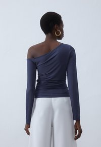 Marinblå långärmad topp med off-shoulder-design, som har samlad textur vid midjan och slätt tyg. Vita byxor syns nedanför.