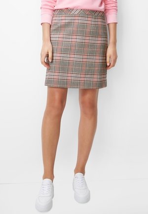 Frau trägt einen hoch taillierten karierten Rock mit roten und schwarzen Linien, ein rosa langärmeliges Oberteil und weiße Plateau-Sneaker vor weißem Hintergrund.