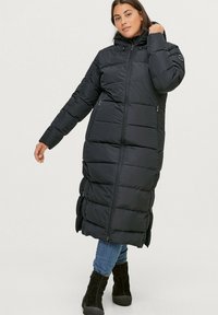 Lange zwarte pufferjas met capuchon, met horizontale stiksels, ritssluiting aan de voorkant en twee zijzakken. Draagt over blauwe jeans en zwarte laarzen.