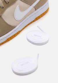 Světlé hnědé tenisky se semišovými a koženými panely, bílým logem Nike a gumovou podrážkou v oranžové barvě. Dvoje bílé ploché tkaničky vedle obuvi.