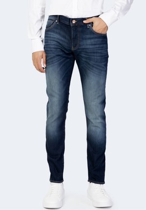 Mann trägt schlanke blaue ausgebleichte Jeans, weißes Hemd und weiße Sneaker, steht mit einer Hand in der Tasche vor einfarbigem Hintergrund.