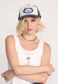 Trucker pet met een licht beige voorkant en een marineblauwe netten achterkant. Voorzien van een ronde klep, een "VOLCOM Authentic" patch en een verstelbare snapback sluiting.