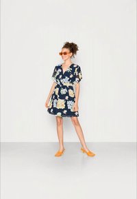 Vestido floral azul-marinho com mangas curtas, decorado com flores azuis claras e amarelas. Combinado com óculos de sol laranja e saltos laranja.