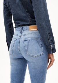 Hellblaue Denim-Jeans mit einer taillierten Passform, zwei Gesäßtaschen und einem quadratischen braunen Etikett am Bund. Glatte Textur und dezente Ausbleichung.