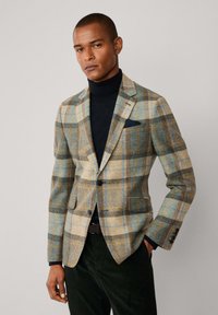 Blazer de cuadros en lana marrón, beige y azul; cuenta con dos botones, solapas con muescas y bolsillos laterales; llevado sobre un cuello de tortuga negro.