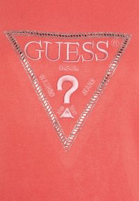 Maglietta color corallo con un logo triangolare con la scritta "GUESS U.S.A." e un bordo decorativo in tono argento. Tessuto morbido con una texture liscia.