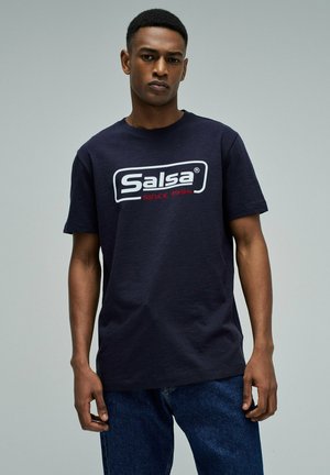 Joven con una camiseta azul marino de Salsa con logo y texto "Desde 1994", combinado con pantalones vaqueros azules, posando frente a un fondo liso.