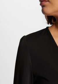 Musta vaate, jossa on V-pääntie ja pehmeät laskostetut olkatiedot, sisältäen sileän tekstuurin ja vartalonmyötäisen muodon.