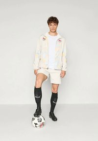 Νεαρός άνδρας με αθλητικά ρούχα, με το ένα πόδι πάνω σε μια μπάλα ποδοσφαίρου, φορώντας ένα ανοιχτό χρωματιστό μπουφάν, σορτς, μαύρες κάλτσες και ποδοσφαιρικά παπούτσια, μπροστά από ουδέτερο φόντο.