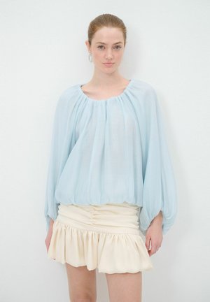 Blusa azzurra chiara con maniche lunghe e arricciate e scollo rotondo; abbinata a una mini gonna crema con volant e arricciature laterali.