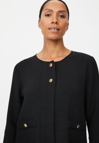 Zwarte gebreide cardigan met ronde halslijn, voorzien van goudkleurige knoopsluitingen en twee steekzakken aan de voorkant.