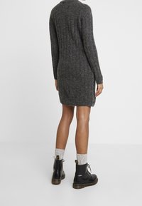 Abito grigio a maglia con maniche lunghe e spacchi laterali, abbinato a stivali neri con lacci e calzini leggeri a maglia. Mostrata la vista da dietro.