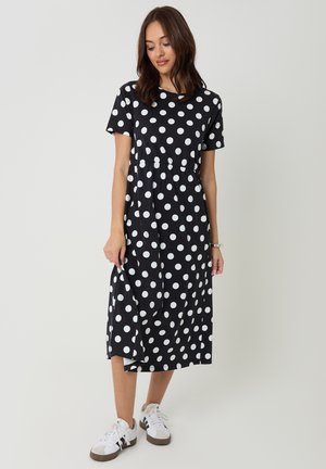 Mujer con vestido midi negro con lunares blancos, mangas cortas y zapatillas blancas, posando contra un fondo liso.