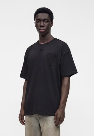 Giovane uomo che indossa una t-shirt nera larga con una tasca sul petto e pantaloni beige chiaro, in piedi davanti a uno sfondo bianco uniforme.