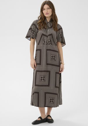 Femme portant une robe mi-longue noire avec des motifs géométriques carrés blancs et des manches courtes volantées, associée à des chaussures plates noires.