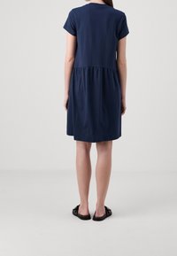 Vrouw die met de rug naar de camera staat, gekleed in een mouwloze, knie-lange marineblauwe jurk en zwarte instap-sandalen op een lichtgrijze achtergrond.