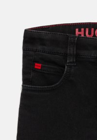 Un jean en denim noir présente une étiquette "HUGO" rouge sur la poche avant, avec des coutures rouges contrastantes le long de la taille. Texture lisse et design ajusté.