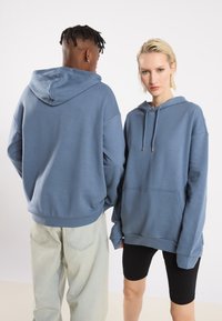 Felpe blu, oversize, con cappuccio e cordini, tasche a marsupio e polsini a coste, indossate da due modelli con stili contrastanti.