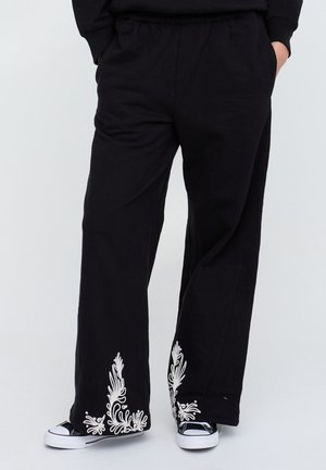 Schwarze, weit geschnittene Hosen mit weißer Blumenstickerei nahe den Säumen, getragen mit schwarzen und weißen Sneakern, Hände in den Taschen.