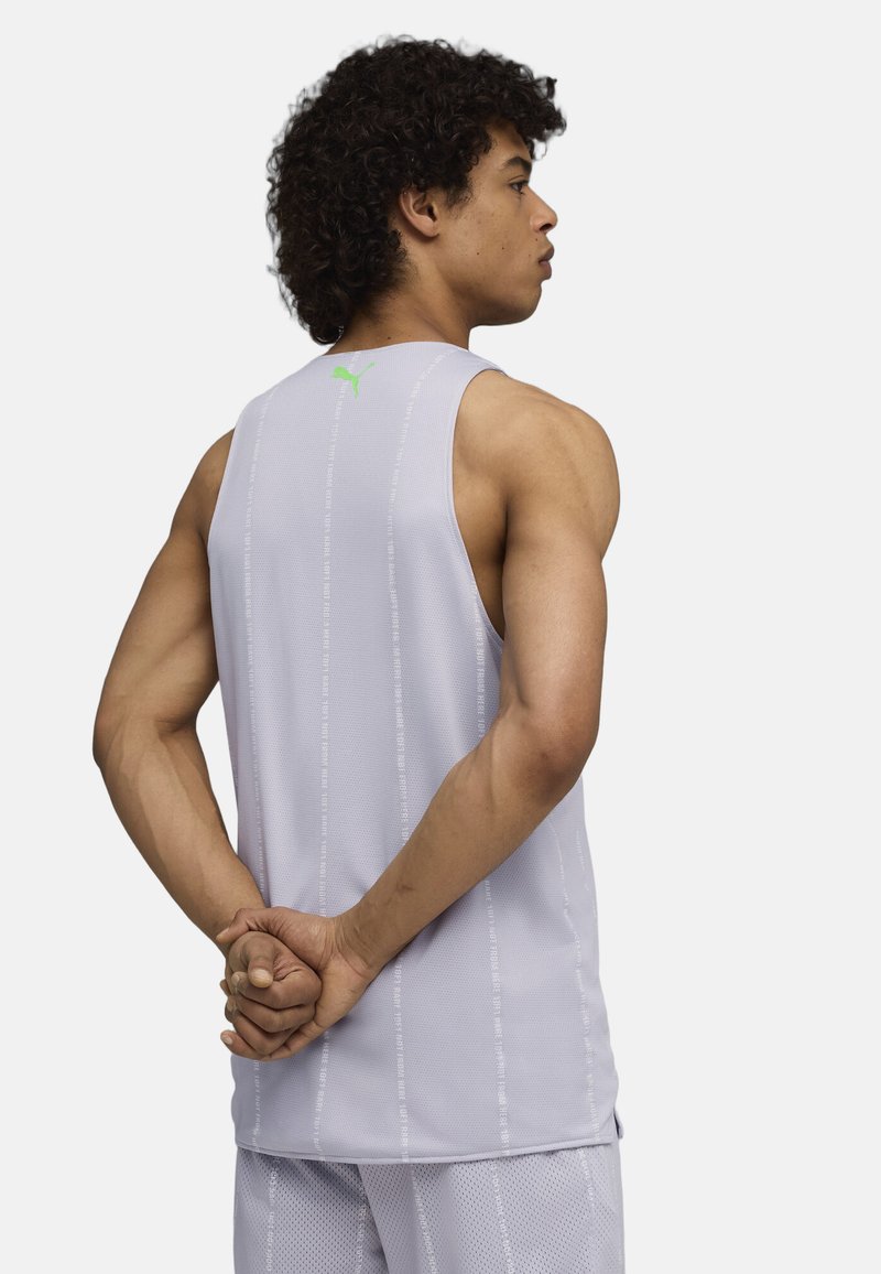 Jeune homme aux cheveux bouclés debout de profil, portant un haut de sport sans manches violet clair et un short assorti, mains jointes dans le dos.
