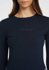 Tamsiai mėlyna ilgomis rankovėmis palaidinė, pagaminta iš raižyto audinio, su subtiliu siuvinėtu "GUESS JEANS" logotipu tos pačios spalvos ant krūtinės.
