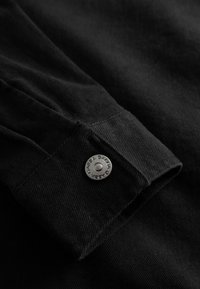 Nærbillede af manchet på en sort denimjakke med en sølvfarvet trykknap, der har indgraveret "GABBA".